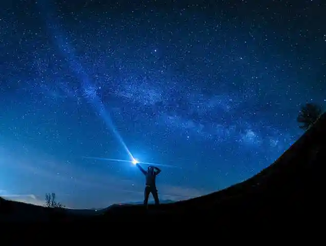 Person on a dark hillside shining a bright flashlight beam toward the Milky Way